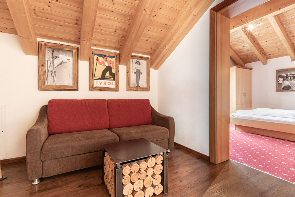 Dieses gemütliche Apartment, Morgensonne für 2-4 Personen in Ladis, verfügt über ein warmes Zimmer mit einem braunen und roten Sofa, einem kleinen Couchtisch aus Holz und gerahmten Bildern an der Wand. Die Holzdecke ergänzt den dunklen Holzboden und führt durch eine offene Tür zu einem hellen Schlafzimmer mit rotem Teppich.