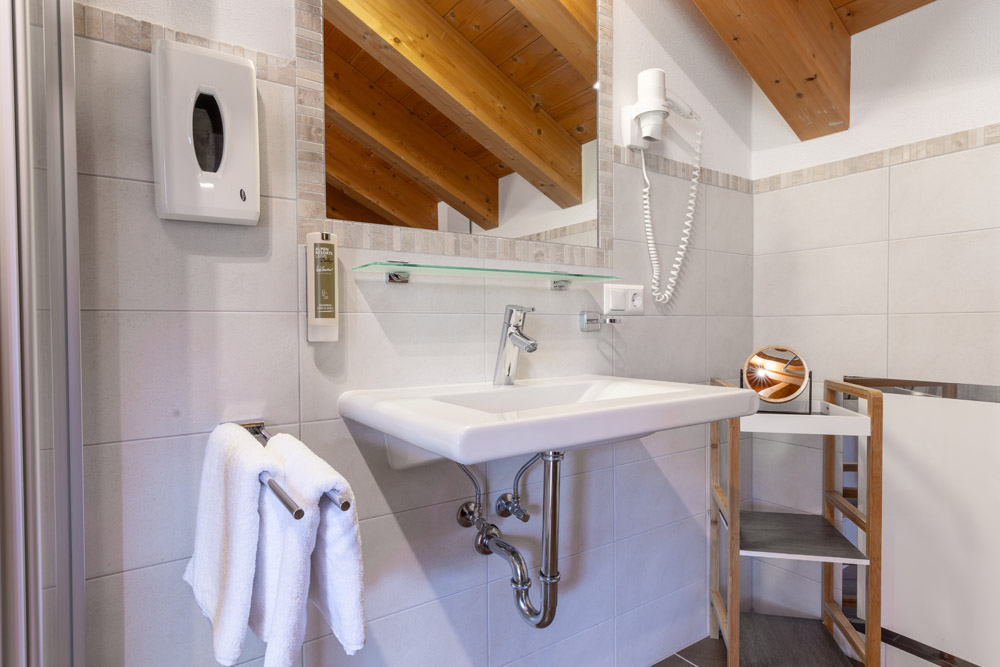 Dieses moderne Badezimmer im charmanten Apartment Morgensonne für 2-4 Personen in Ladis verfügt über ein elegantes weißes Waschbecken und einen großen Spiegel. Ein flauschiges weißes Handtuch hängt an einem Ständer und ergänzt den montierten Seifenspender und den Haartrockner. Die Holzbalken an der Decke verleihen den hellgrau gefliesten Wänden einen Hauch von Wärme.