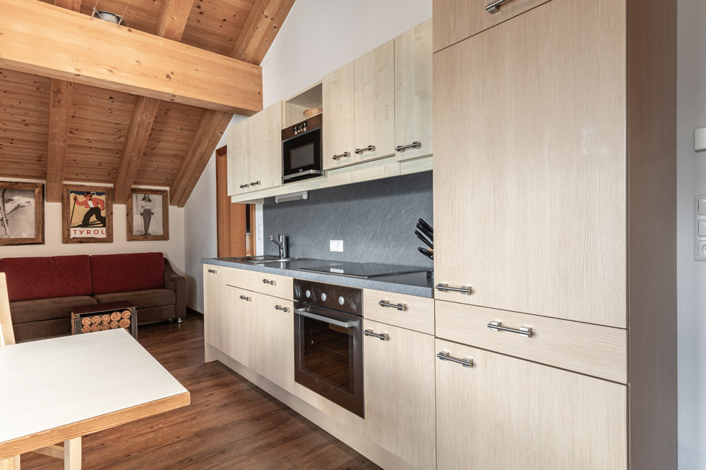 Eine gemütliche Küche im Apartment Morgensonne für 2-4 Personen in Ladis verfügt über helle Holzschränke und eine dunkle Arbeitsplatte, mit Backofen, Herd und wandmontierter Mikrowelle. Ein rotes Sofa und gestapeltes Brennholz stehen unter einer schrägen Holzdecke, die mit alpiner Kunst geschmückt ist.