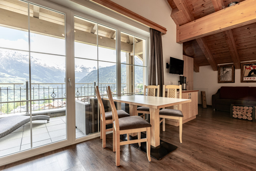 Dieser gemütliche alpine Wohnbereich im Apartment Morgensonne für 2-4 Personen in Ladis verfügt über Holzböden und Deckenbalken. Ein Esstisch mit vier Stühlen steht neben großen Glastüren, die auf einen Balkon mit herrlichem Bergblick führen, während ein Fernseher und ein Sofa in der Ecke die warme Atmosphäre verstärken.