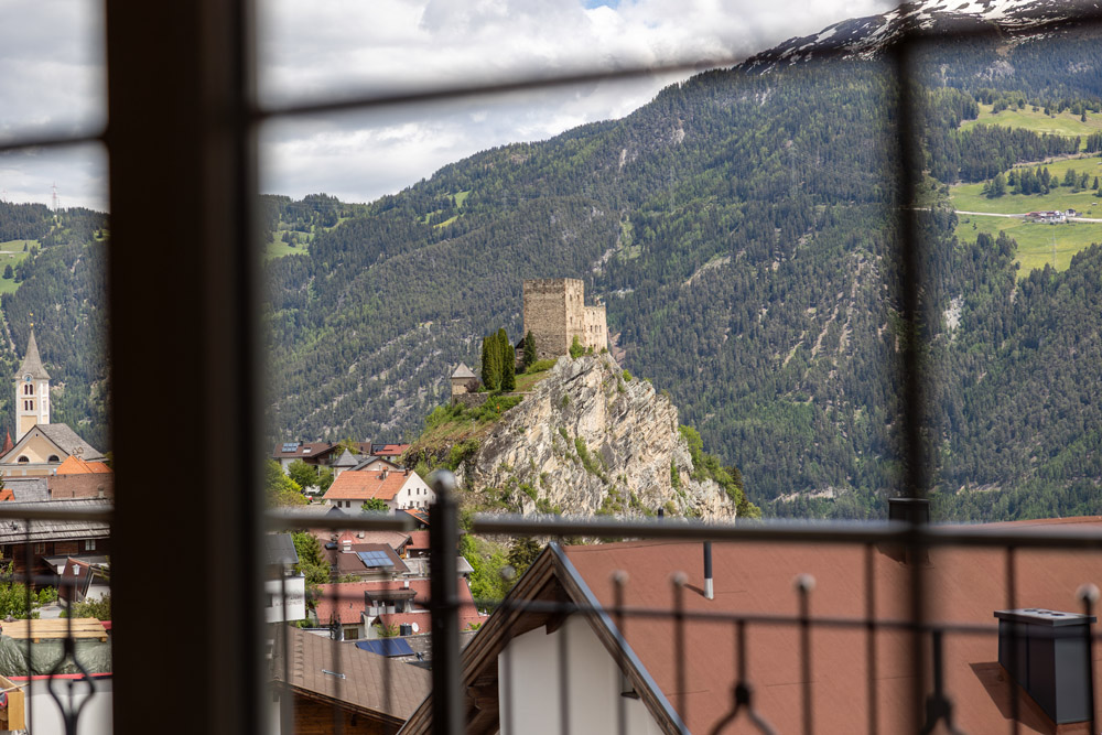 Wenn man durch das Fenster einer mittelalterlichen Steinburg auf einem felsigen Hügel blickt, umgeben von einem Dorf und üppigen grünen Bergen, erinnert man sich an ein Apartment Morgensonne für 2-4 Personen in Ladis. Der Fensterrahmen verdeckt teilweise die Szene, während in der Ferne majestätisch schneebedeckte Gipfel aufragen.