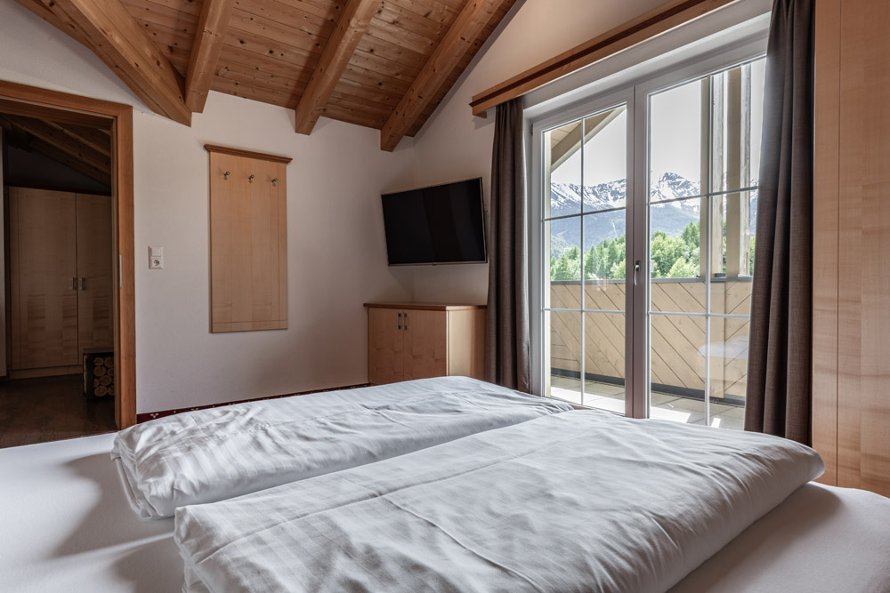 Das gemütliche Schlafzimmer im Apartment Morgensonne für 2-4 Personen in Ladis verfügt über eine schräge Holzdecke, ein Doppelbett und einen Flachbildfernseher. Ein großes Fenster bietet einen atemberaubenden Blick auf schneebedeckte Berge, während sanftes Sonnenlicht die hellen Holzmöbel und die weiße Bettwäsche durchflutet.