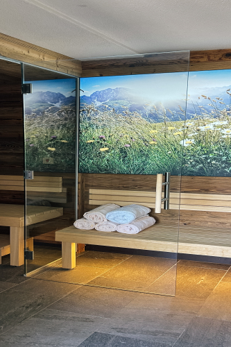 Ein moderner Sauna-Innenraum im Stil eines Familienchalets für 4-6 Personen in Ladis, mit Holzbänken und einer eleganten Glastür. Gefaltete Handtücher liegen ordentlich auf der Bank, während die Wand ein lebendiges Landschaftsbild mit Bergen und Wildblumen zeigt. Der Boden besteht aus eleganten Steinfliesen.