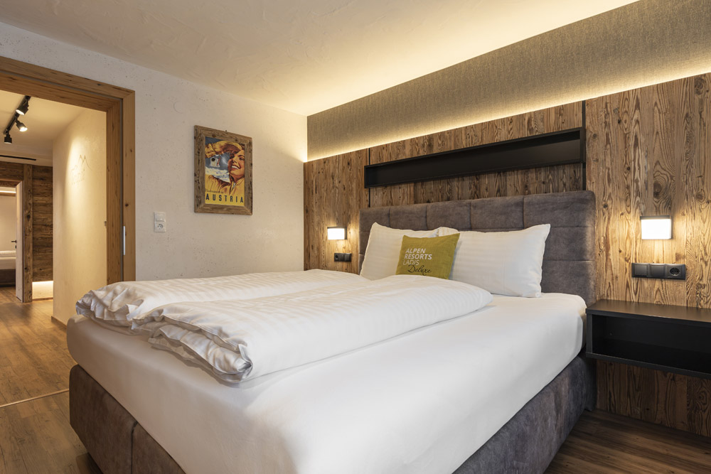 Ein gemütliches Hotelzimmer, das an ein Familienchalet erinnert, mit einem Doppelbett mit weißer Bettwäsche und einem grünen Zierkissen. Die Wände sind mit Holzvertäfelungen und einem Vintage-Poster mit Österreich-Motiv geschmückt, das den Charme von Ladis heraufbeschwört. Sanfte Beleuchtung schafft eine warme Atmosphäre in dem einladenden Raum.