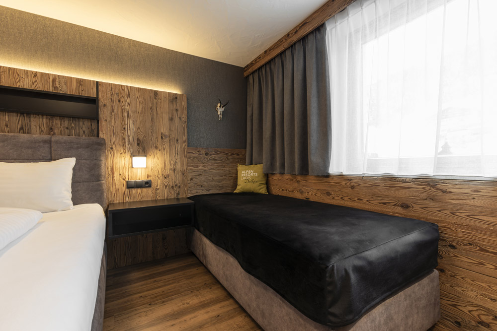 Dieses Familienchalet in Ladis bietet bequem Platz für 4-6 Personen. Das gemütliche, mit Holzvertäfelung ausgestattete Zimmer verfügt über ein Doppel- und ein Einzelbett. Über dem Kissen neben dem Fenster, das von transparenten, dunklen Vorhängen eingerahmt wird, thront ein dekorativer Stierkopf.