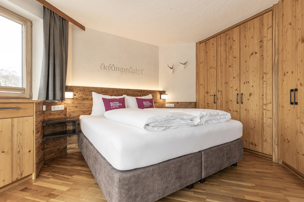 Gemütliches Schlafzimmer in einem Familienchalet für 4–6 Personen in Ladis, mit einem großen Bett mit burgunderfarbenen Kissen und weißer Bettwäsche. Die Holzböden und Kiefernholzschränke sorgen für Wärme, während natürliches Licht durch das Fenster strömt. Dekorative Geweihe schmücken die Wände und sorgen für eine charmante Note.