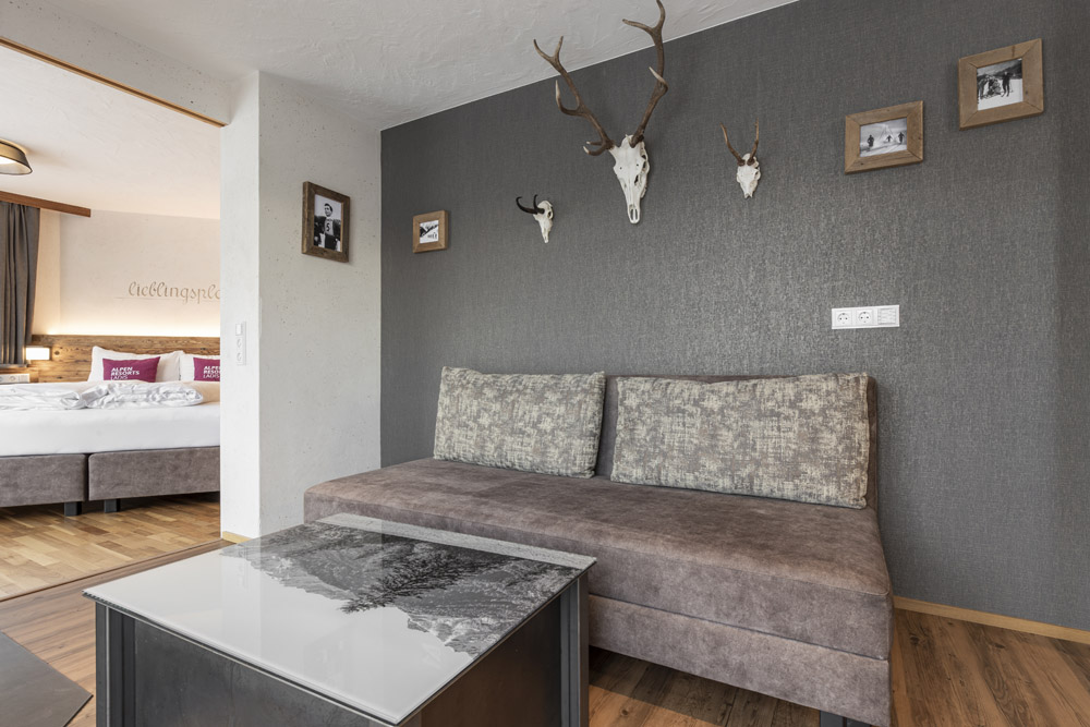 Modernes Hotelzimmer in einem Familienchalet für 4-6 Personen in Ladis, mit einem grauen Sofa und dekorativen Geweihen an einer strukturierten dunklen Wand. Gerahmte Fotos begleiten die Geweihe, im Hintergrund sind ein ordentlich gemachtes Bett und ein hölzernes Kopfteil zu sehen. Im Vordergrund steht ein Couchtisch.