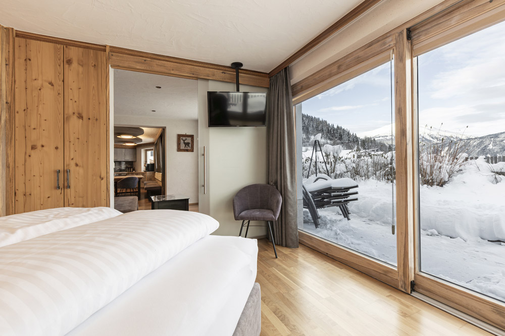 Ein gemütliches Familienchalet für 4-6 Personen in Ladis, mit einem Schlafzimmer mit weißer Bettwäsche, Holzwänden und einem an der Wand montierten Fernseher. Große Fenster geben den Blick auf die verschneite Landschaft und die Berge draußen frei. Ein grauer Stuhl steht am Fenster und bietet Komfort, während Sie hinausblicken oder in ein anderes Zimmer spähen.