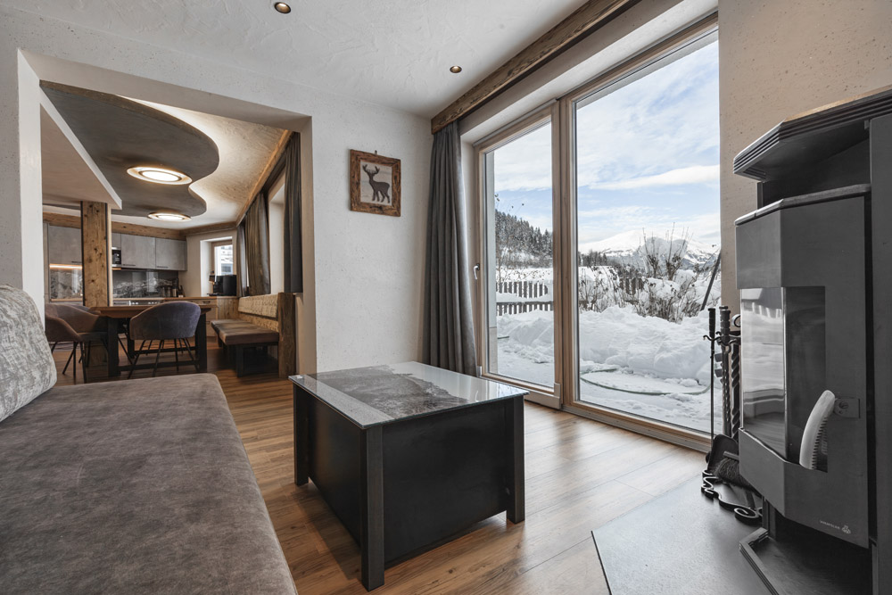 Modernes Wohnzimmer in einem Familienchalet für 4-6 Personen in Ladis, mit einem großen Fenster mit Blick auf die verschneite Landschaft. Der Raum verfügt über ein graues Sofa, einen Couchtisch aus Glas und einen eleganten Kamin. Im offenen Bereich sind ein Esstisch und Stühle aus Holz vor weißen Wänden mit Holzakzenten zu sehen.