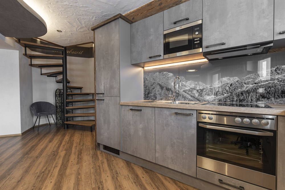Dieses Familienchalet in Ladis bietet eine moderne Küche mit hellen Holzschränken, einem Backofen unter einer Mikrowelle und einer markanten schwarz-weißen Küchenrückwand mit Bergmotiv. Holzböden und freiliegende Balken ergänzen den Raum, während eine Wendeltreppe nach oben führt und Platz für 4-6 Personen bietet.
