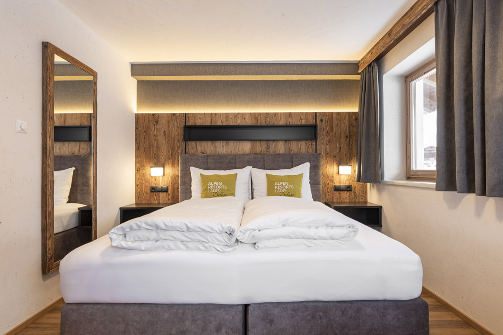 Ein gemütliches Hotelzimmer, das an ein Familienchalet für 4-6 Personen in Ladis erinnert, verfügt über ein ordentlich gemachtes Doppelbett mit weißer Bettwäsche und gelben Kissen. Das Zimmer verfügt über ein hölzernes Kopfteil, einen großen Spiegel an der linken Wand und ein Fenster mit geschlossenen Vorhängen auf der rechten Seite.