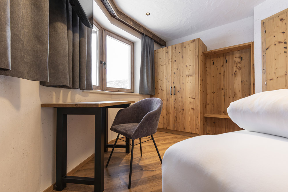 Ein gemütliches Zimmer in einem Familienchalet für 4-6 Personen in Ladis mit Holzboden und -möbeln, darunter ein Kleiderschrank, ein Schreibtisch und ein Stuhl. Ein Bett mit weißer Bettwäsche ist teilweise sichtbar. Weiches Licht fällt durch die beiden mit dunklen Vorhängen behangenen Fenster.