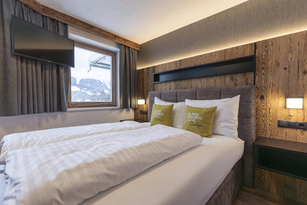 Gemütliches Hotelzimmer im Stil eines Familienchalets für 4-6 Personen in Ladis, ausgestattet mit einem Doppelbett mit weißer Bettwäsche und grünen Kissen. An der holzgetäfelten Wand ist ein Flachbildfernseher angebracht und ein Fenster bietet einen Blick auf die schneebedeckten Berge, der durch warmes Licht für eine einladende Atmosphäre sorgt.