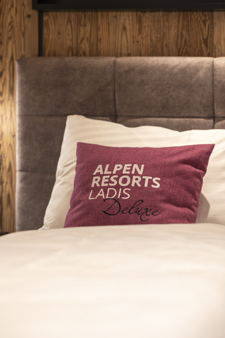 Ein gemütliches Familienchalet für 4-6 Personen in Ladis verfügt über ein Bett mit einem weichen Kopfteil und frischen weißen Laken. Darauf liegt ein lila Kissen mit der Aufschrift „ALPEN RESORTS LADIS Deluxe“, alles vor einer warmen Holzwandkulisse.