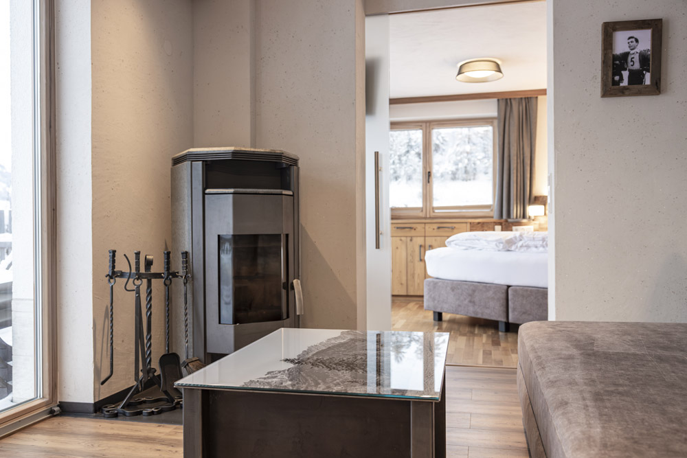 Gemütlicher Wohnbereich in einem Familienchalet für 4-6 Personen in Ladis mit einem Couchtisch mit Glasplatte, einem Holzofen mit Werkzeug daneben und einem gerahmten Foto an der Wand. Eine Öffnung führt zu einem Schlafzimmer mit einem Bett und Holzschränken, während große Fenster den Raum erhellen.