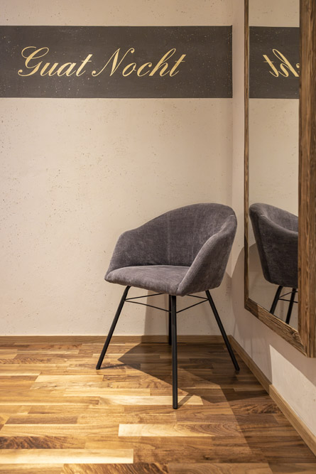 Ein modernes Zimmer in unserem Familienchalet für 4-6 Personen in Ladis verfügt über einen grauen Polsterstuhl auf einem Holzboden. Ein großer Spiegel reflektiert einen Teil des Stuhls, während ein dekorativer Streifen die Wand mit der eleganten goldenen Aufschrift „Guat Nocht“ ziert.