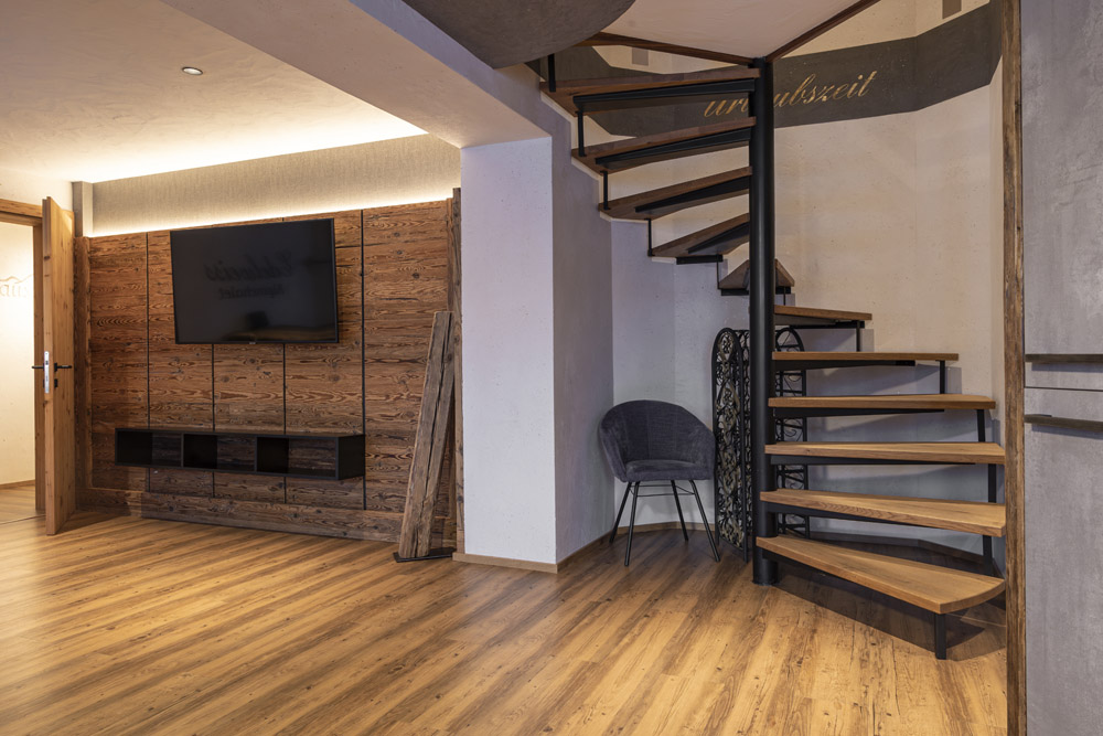 Ein modernes Wohnzimmer mit Holzboden und Wendeltreppe verströmt den Charme eines Familienchalets für 4-6 Personen in Ladis. Die linke Wand verfügt über einen großen Fernseher auf einer strukturierten Holzplatte, während ein grauer Stuhl neben der Treppe eine gemütliche Ecke schafft, in der rustikale und moderne Atmosphäre nahtlos miteinander verschmelzen.