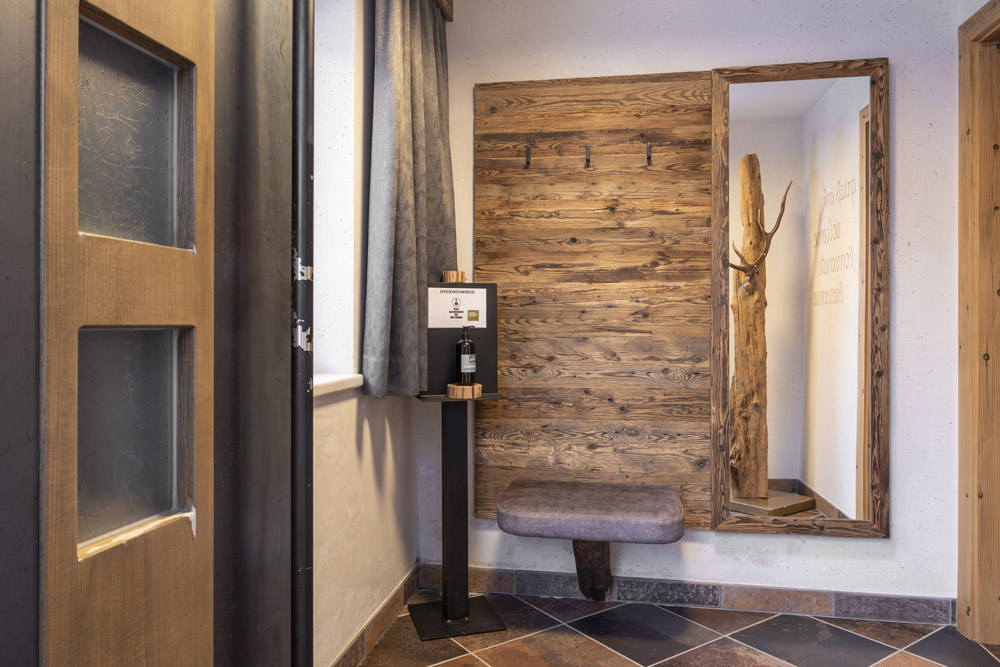 Ein rustikales Familien-Chalet-Zimmer für 4-6 Personen in Ladis mit Holzwänden, einem großen Spiegel und einer Garderobe aus Holz. Eine Desinfektionsstation befindet sich links neben einer gepolsterten Bank. Natürliches Licht fällt durch ein teilweise sichtbares Fenster auf den gefliesten Boden.