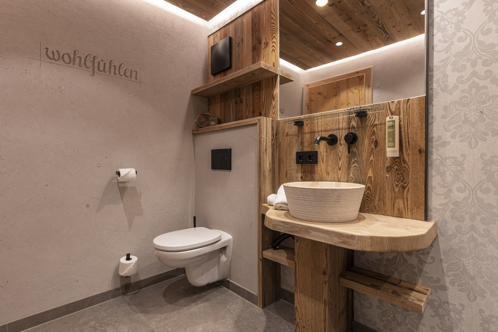 Modernes Badezimmer mit rustikalen Holzakzenten. Verfügt über eine wandmontierte Toilette, ein rundes Steinwaschbecken auf einem Waschtisch aus Naturholz und dezente Beleuchtung. Die Wände sind strukturiert und tragen den Text „Wohlfühlen“. Graue Fliesenböden und ein gemusterter Wandabschnitt sind ebenfalls sichtbar.