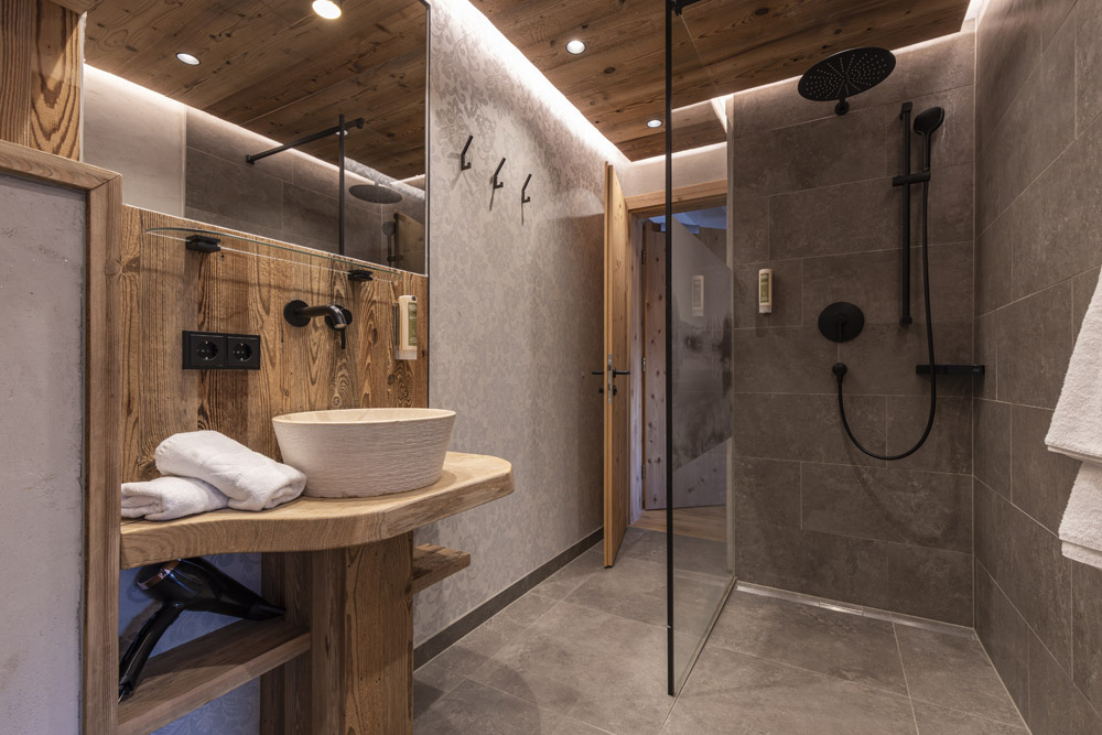 Ein modernes Badezimmer mit rustikaler Ästhetik verfügt über eine Holzdecke und ein Waschbecken. Ein erhöhtes Waschbecken wird von einem wandmontierten Wasserhahn begleitet. Links sind Handtücher und rechts ist ein Regenduschkopf und eine Glaskabine.
