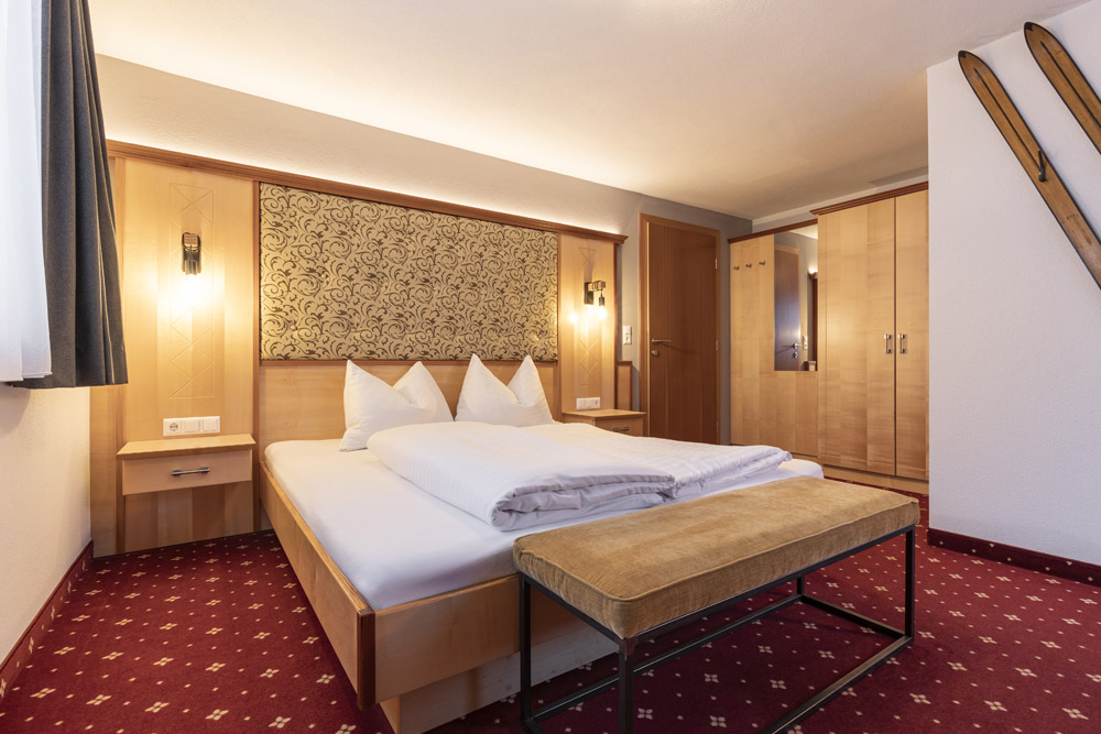 Genießen Sie ein charmantes Hotelzimmer, das sich ideal für einen Urlaub zu Zweit in Serfaus-Fiss-Ladis eignet. Es verfügt über ein ordentlich gemachtes Doppelbett, Holzmöbel und ein gemustertes Kopfteil. Skier zieren kunstvoll die Wand und der rote Teppich des Zimmers verleiht mit seinem einfachen Motiv Wärme.