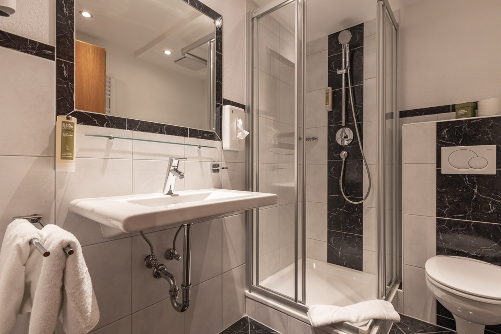 Dieses moderne Badezimmer, perfekt für einen romantischen Urlaub zu Zweit in Serfaus-Fiss-Ladis, verfügt über eine Dusche mit Glaskabine, schwarz-weißes Fliesendesign, ein wandmontiertes Waschbecken mit Spiegel darüber, einen Handtuchhalter mit frischen weißen Handtüchern und wird durch elegante Einbauleuchten beleuchtet.