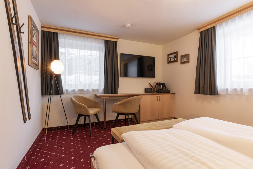 Genießen Sie für Ihren „Urlaub zu Zweit in Serfaus-Fiss-Ladis“ ein gemütliches Hotelzimmer mit Doppelbett, rotem Teppich und weißen Wänden. Es gibt einen Flachbildfernseher auf einem Holzschrank, zwei Stühle an einem Schreibtisch, Vintage-Skier an der Wand und große Fenster mit grauen Vorhängen, die natürliches Licht hereinlassen.
