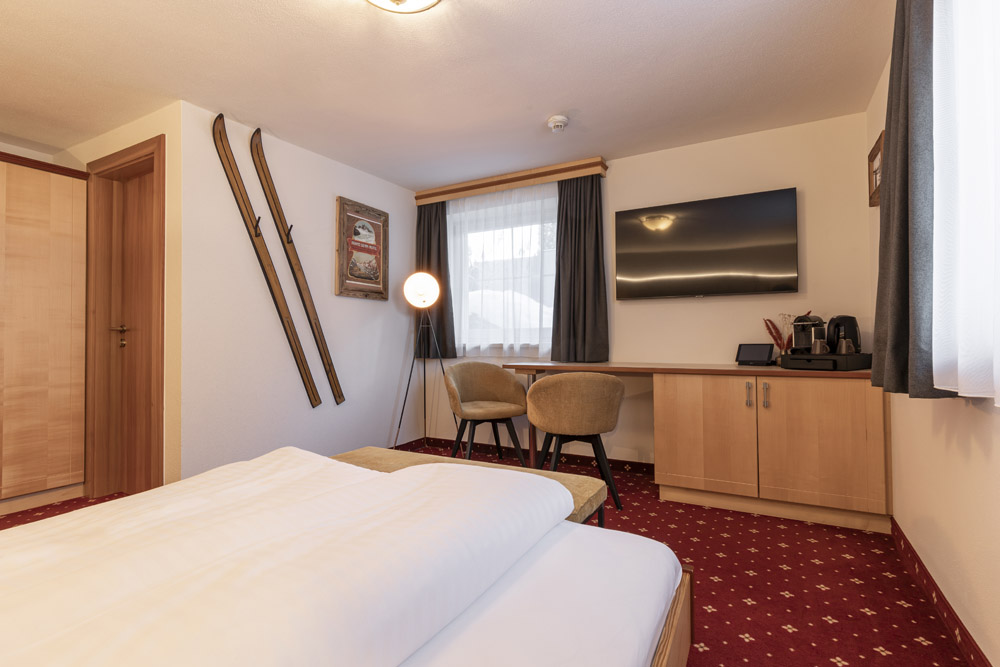 Ein gemütliches Hotelzimmer perfekt für Urlaub zu Zweit in Serfaus-Fiss-Ladis, mit einem Bett im Vordergrund, Vintage-Skiern an der Wand, einem großen Flachbildfernseher, einem Schreibtisch für zwei und einer modernen Lampe. Der rot gemusterte Teppich ergänzt das Fenster mit Vorhängen.