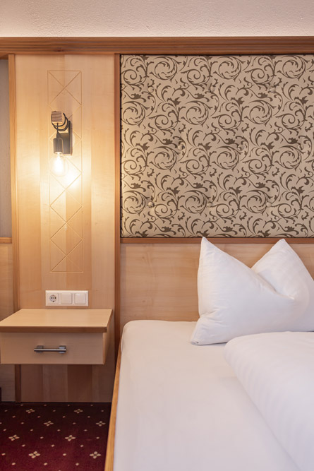 Eine gemütliche Hotelzimmerecke, perfekt für einen Urlaub zu zweit in Serfaus-Fiss-Ladis, mit einem Bett mit frischen weißen Kissen und Bettwäsche. Ein sanfter Schein der Wandlampe neben dem Bett erwärmt den Raum über einem kleinen Nachttisch aus Holz. Das Zimmer verfügt über eine gemusterte beige-braune Wandverkleidung, ergänzt durch einen rotgemusterten Teppich.