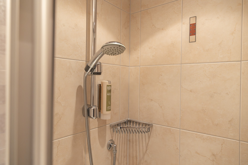 Eine moderne Dusche in einem Apartment für 2-3 Personen in Serfaus-Fiss-Ladis verfügt über beige geflieste Wände, eine Handbrause und einen montierten Seifenspender. Ein Metalldrahtkorb bietet Stauraum, während eine dekorative rechteckige Fliese für eine elegante Note sorgt.