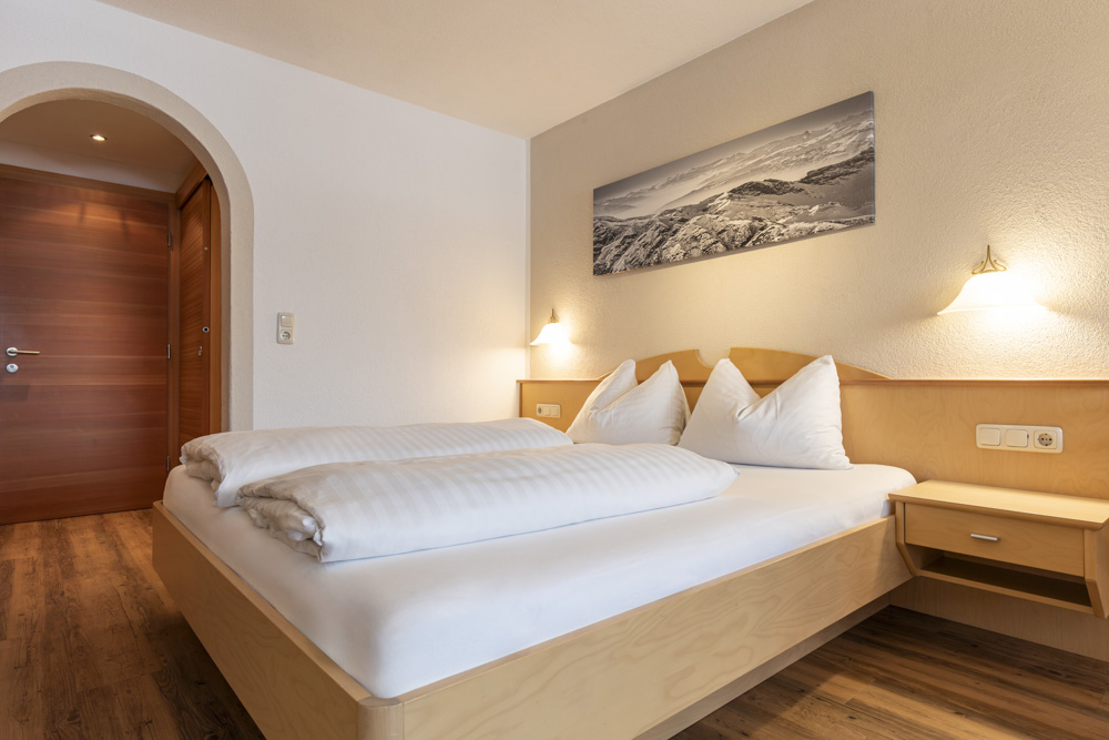 Ein gemütliches Schlafzimmer in einem Apartment für 2-3 Personen in Serfaus-Fiss-Ladis verfügt über ein großes Bett mit weißer Bettwäsche und Holzmöbeln. Warmes Licht und ein schwarz-weißes Landschaftsfoto verstärken die einladende Atmosphäre, während im Hintergrund eine Holztür zu sehen ist.