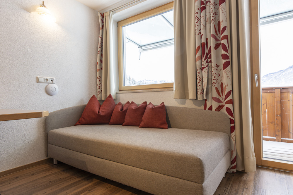 Ein gemütliches Apartment für 2-3 Personen in Serfaus-Fiss-Ladis verfügt über ein beiges Sofa mit fünf roten Kissen. Es hat Holzboden, ein großes Fenster mit geblümten Vorhängen und Blick auf den Holzbalkon. Sanftes Licht erhellt sanft die weißen Wände.