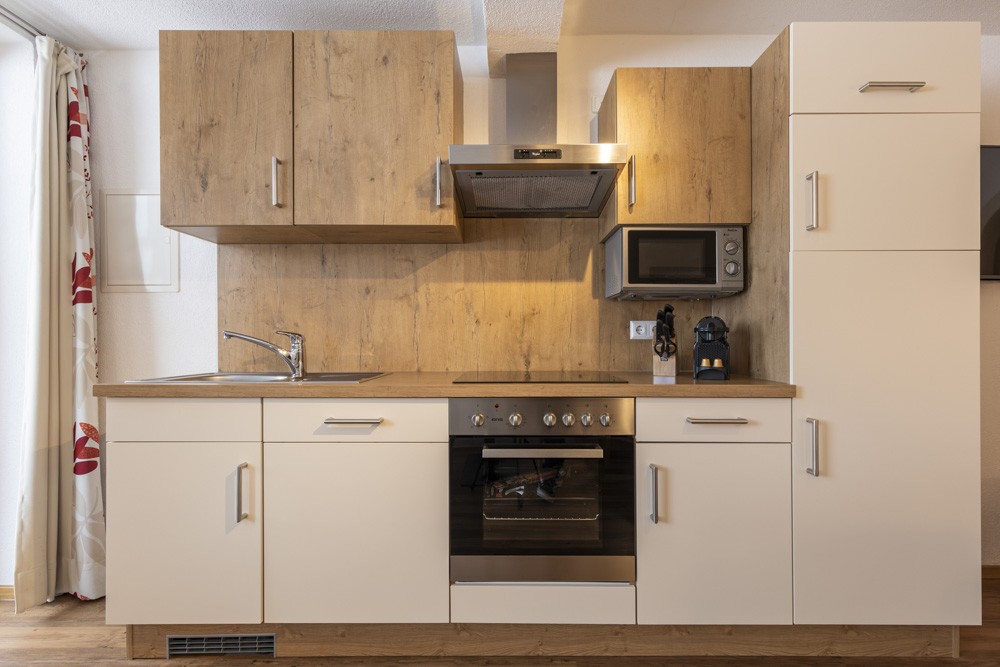 Diese moderne Küche in einem Apartment für 2-3 Personen in Serfaus-Fiss-Ladis verfügt über Holz- und weiße Schränke, eine Edelstahlspüle, einen Elektroherd, einen Backofen, eine Dunstabzugshaube, eine Mikrowelle und eine Kaffeemaschine. Ein großer weißer Kühlschrank steht rechts unter einem Fenster mit gemusterten Vorhängen.