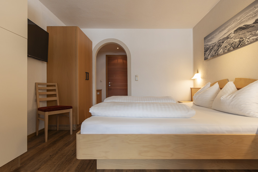 Ein minimalistisches Hotelzimmer, das einem Apartment für 2-3 Personen in Serfaus-Fiss-Ladis ähnelt, verfügt über ein ordentlich gemachtes Doppelbett mit weißer Bettwäsche, einen an der Wand montierten Fernseher, einen Holzschrank und einen einzelnen Stuhl. An der Wand hängt ein Landschaftsbild, und sanfte Innenbeleuchtung verleiht dem Raum Wärme.
