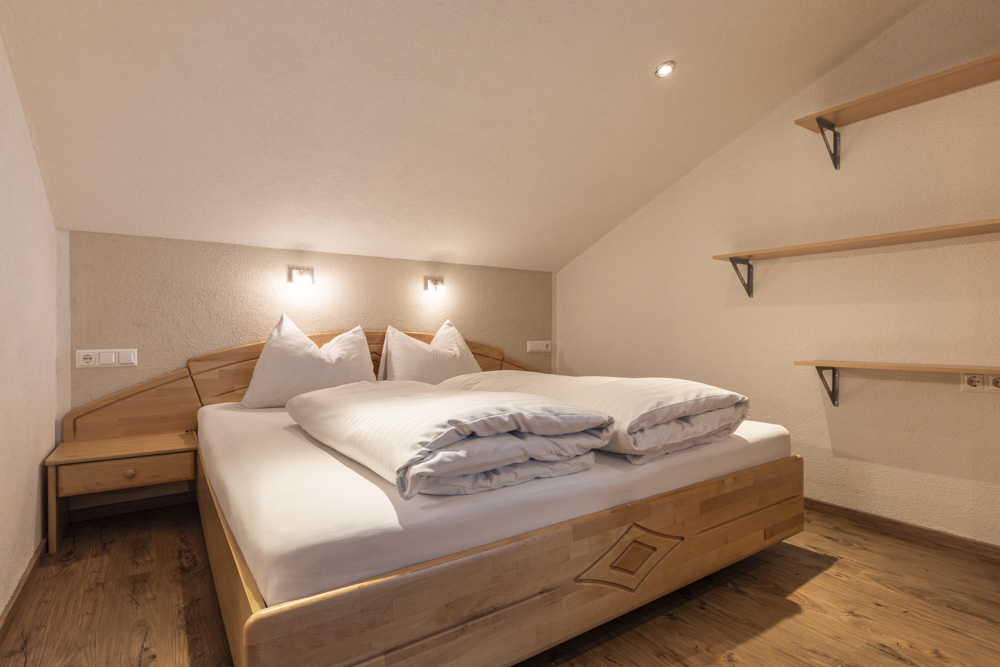Ein gemütliches Schlafzimmer in einem Apartment für 2-3 Personen in Serfaus-Fiss-Ladis verfügt über ein Doppelbett mit weißer Bettwäsche und einem hölzernen Kopfteil. Flankiert von Nachttischen und beheizt durch Wandlampen, bietet der Raum Hartholzböden und leere Regale unter einer Dachschräge.