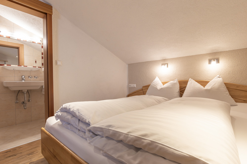Gemütliches Schlafzimmer im Dachgeschoss eines Apartments für 2-3 Personen in Serfaus-Fiss-Ladis mit einem sauber gemachten Doppelbett mit weißer Bettwäsche. Warmes Licht sorgt für eine einladende Atmosphäre. Eine offene Tür führt zu einem kleinen Badezimmer mit Waschbecken und großem Spiegel.