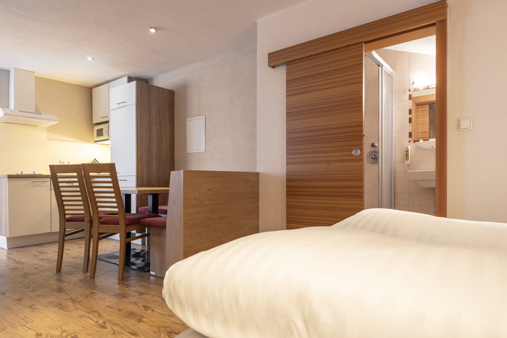 Modernes Studio-Apartment mit Holzboden, ideal für 2-3 Personen in Serfaus-Fiss-Ladis. Es verfügt über eine kleine Küche mit weißen Schränken, einen Esstisch mit Holzstühlen und eine teilweise geöffnete Schiebetür, die zu einem kompakten Badezimmer führt. Im Vordergrund steht ein Bett mit weißer Bettwäsche.