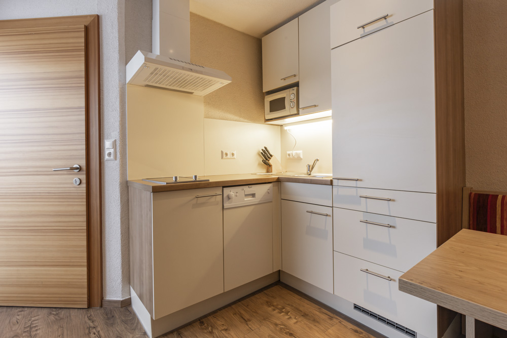 Eine kompakte, moderne Küche, die auf ein Apartment für 2-3 Personen in Serfaus-Fiss-Ladis zugeschnitten ist, verfügt über weiße Schränke, einen Herd, eine Spüle und einen Geschirrspüler. Holzakzente auf dem Boden und den Schrankrahmen sorgen für Wärme, ergänzt durch eine kleine Arbeitsplatte und eine Holztür auf der linken Seite.