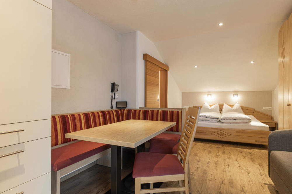 Gemütliches Apartment für 2-3 Personen in Serfaus-Fiss-Ladis mit einem Zimmer im Hotelstil mit einem Holztisch und roten gepolsterten Stühlen, einer Sitzecke und einem Doppelbett mit weißer Bettwäsche. Genießen Sie Holzböden, ein kleines Wandtelefon und warmes Licht für zusätzlichen Komfort.