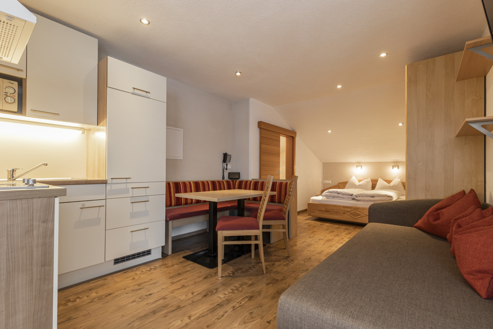 Gemütliches Studio-Apartment für 2-3 Personen in Serfaus-Fiss-Ladis, mit einem kleinen Küchenbereich mit Holzschränken, einer Sitzecke mit einer rot gestreiften Bank und Holzstühlen, einer grauen Couch mit orangefarbenen Kissen und einem Bett mit weißer Bettwäsche. Der Holzboden verleiht dem Raum Wärme.