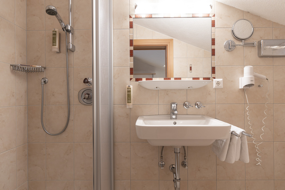 Das kompakte Badezimmer im Apartment für 2-3 Personen in Serfaus-Fiss-Ladis verfügt über beige geflieste Wände, eine ebenerdige Dusche mit Metallregal, ein weißes Waschbecken unter einem rechteckigen Spiegel, einen montierten Haartrockner und einen runden Rasierspiegel mit Vergrößerung. Für mehr Komfort hängt ein Handtuch über dem Waschbecken.