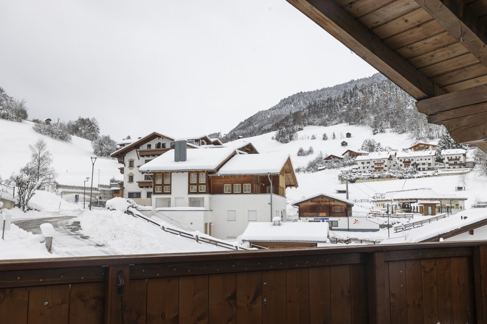 Ein schneebedecktes Alpendorf mit Holzhäusern vor einer Bergkulisse setzt den Rahmen. Ein nahe gelegenes Apartment für 2-3 Personen in Serfaus-Fiss-Ladis bietet gemütliche Unterkunft. Der bedeckte Himmel und die schneebedeckten Bäume verstärken die winterliche Atmosphäre, ein Holzgeländer rahmt die Aussicht ein.