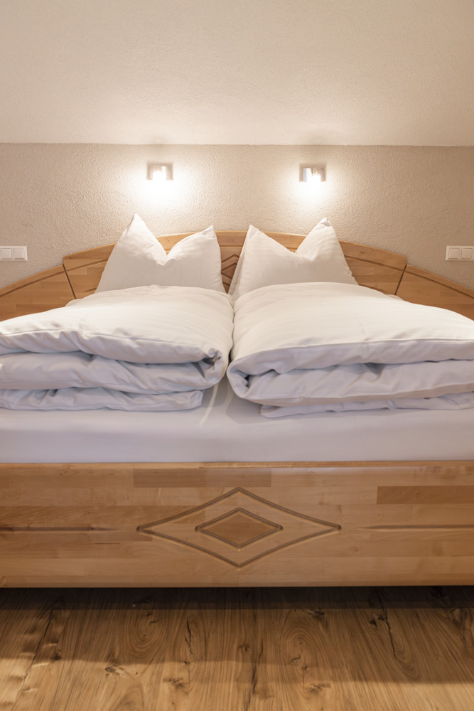 Ein ordentlich gemachtes Doppelbett mit weißen Kissen und Bettwäsche steht in einem warm beleuchteten Raum, perfekt für ein Apartment für 2-3 Personen in Serfaus-Fiss-Ladis. Das hölzerne Kopfteil weist ein geometrisches Design auf. An der Wand über dem Bett sind zwei kleine Lampen angebracht, die den Holzboden ergänzen.