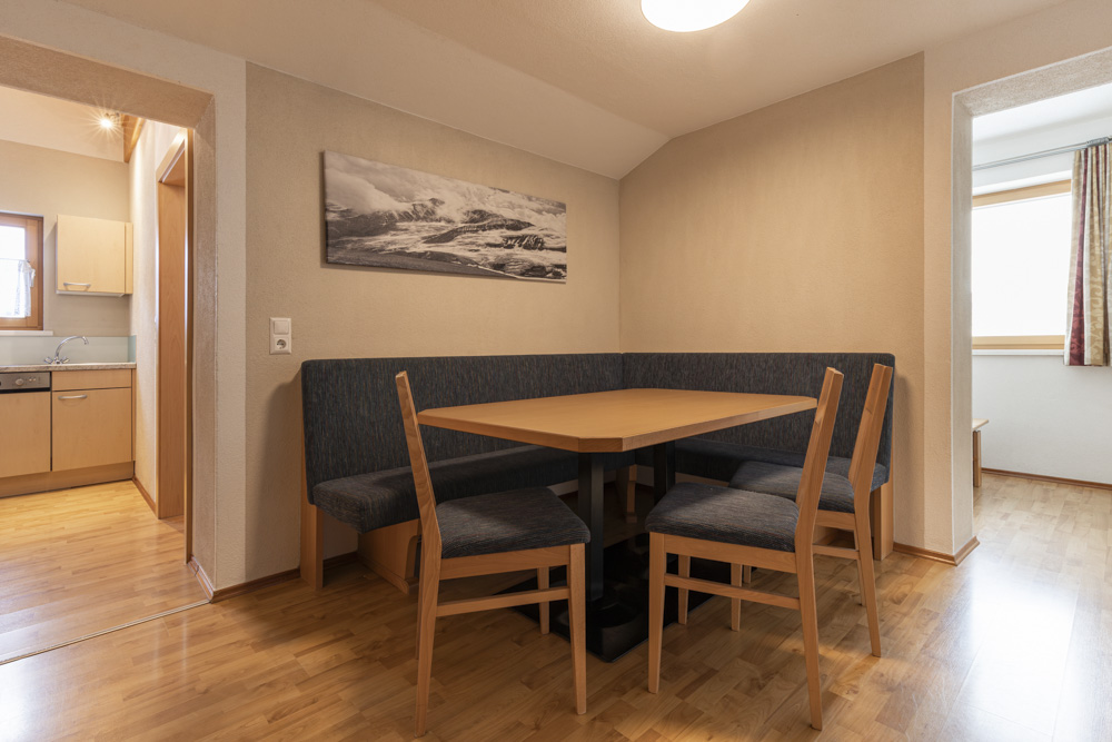 Eck-Essbereich in einem Apartment für 4-6 Personen in Serfaus-Fiss-Ladis, mit einem Holztisch mit Sitzbank und drei Stühlen. Heller Holzboden ergänzt das schwarz-weiße Berglandschaftsbild, das über der Bank hängt, mit Blick durch die Türen in die angrenzenden Räume.