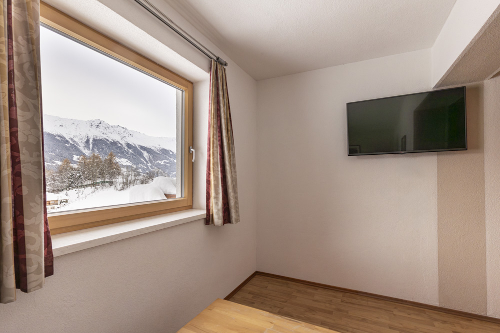 Ein minimalistisches Zimmer in einem Apartment für 4-6 Personen in Serfaus-Fiss-Ladis verfügt über einen Holzboden, einen an der Wand montierten Fernseher und ein großes Fenster mit Blick auf eine schneebedeckte Berglandschaft mit Bäumen. Helle Vorhänge rahmen die atemberaubende Aussicht ein.