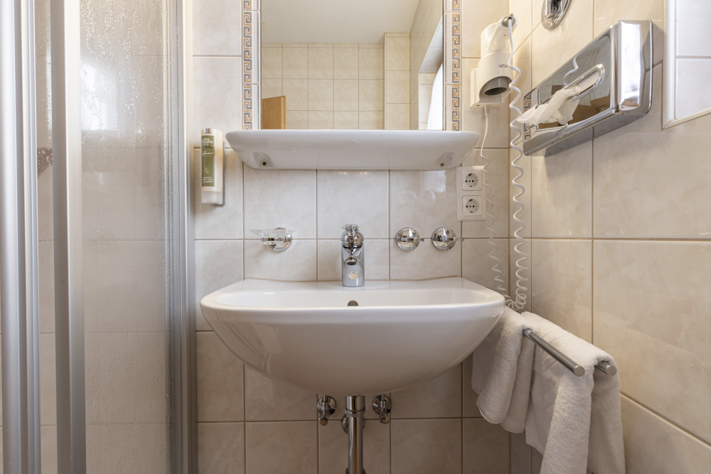 Ein kleines Badezimmer in einem Apartment für 4-6 Personen in Serfaus-Fiss-Ladis verfügt über beige geflieste Wände, ein weißes Waschbecken unter einem Wandregal und zwei gefaltete Handtücher auf einer Stange unter dem Waschbecken. Ein montierter Haartrockner und eine teilweise sichtbare Duschkabine vervollständigen diesen gemütlichen Raum.
