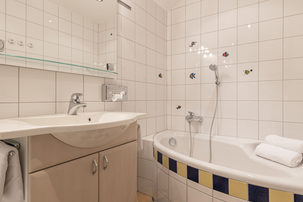Das charmante Badezimmer im Apartment für 4-6 Personen Serfaus-Fiss-Ladis besticht durch weiß geflieste Wände mit bezaubernden Fischdekorationen. Es verfügt über ein Waschbecken mit Spiegel und Ablage, ein Handtuch links und eine Eckbadewanne mit Duschkopf, die von zwei aufgerollten weißen Handtüchern geschmückt wird.