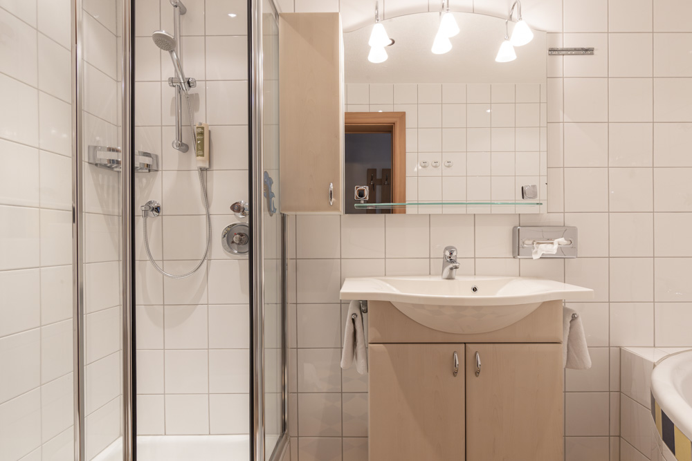 Dieses moderne Badezimmer, Teil eines Apartments für 4-6 Personen in Serfaus-Fiss-Ladis, verfügt über weiß geflieste Wände und eine verglaste Dusche. Ein hölzerner Waschtisch mit weißem Waschbecken befindet sich unter einem großen Spiegel, der von drei Glühbirnen beleuchtet wird. An der Seite des Schranks hängt ordentlich ein Handtuch.