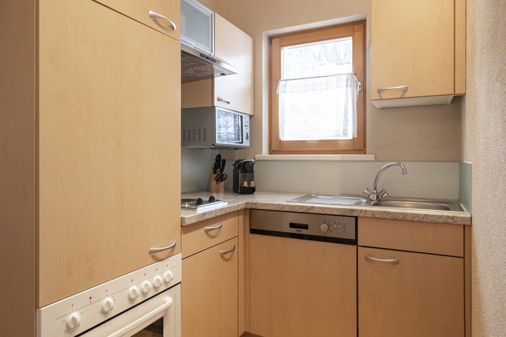 Eine kompakte Küche in einem Apartment für 4-6 Personen Serfaus-Fiss-Ladis verfügt über helle Holzschränke, einen modernen Herd und eine Edelstahlspüle. Ein Fenster mit Spitzenvorhang sorgt für Tageslicht. Eine Mikrowelle, eine kleine Arbeitsfläche und ein Messerset vervollständigen den gemütlichen Kochbereich.
