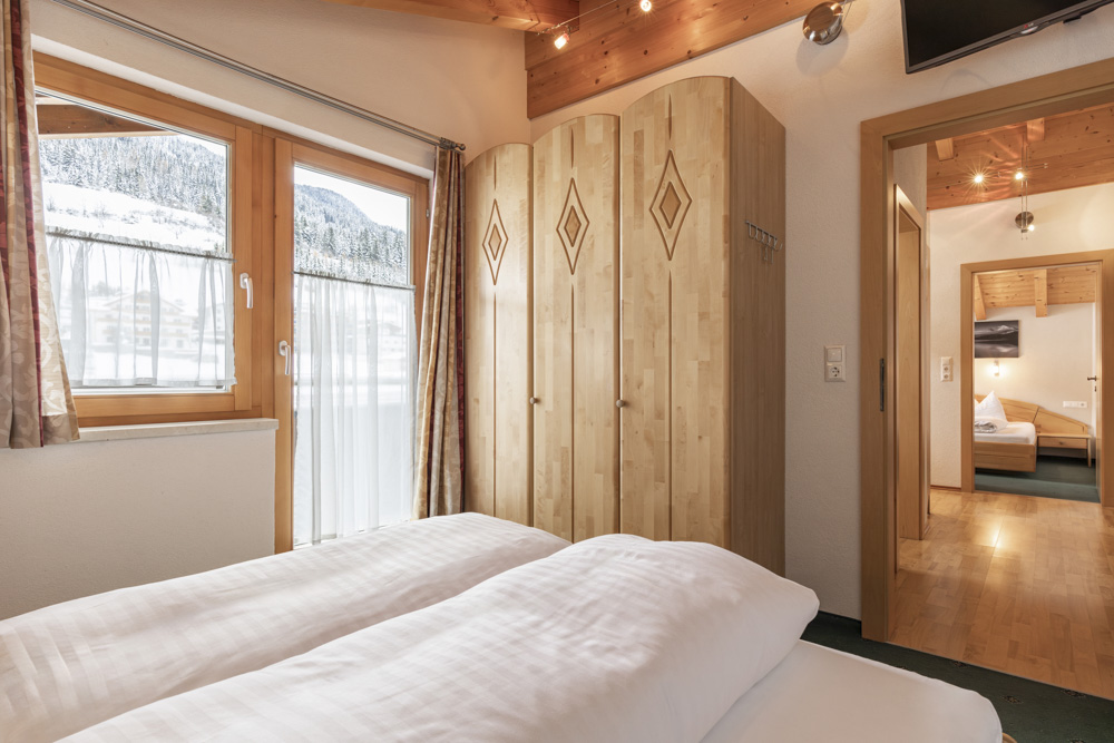 Dieses gemütliche Apartment für 4-6 Personen in Serfaus-Fiss-Ladis bietet ein charmantes Schlafzimmer mit einem Doppelbett mit weißer Bettwäsche, hellen Holzpaneelen und einem Kleiderschrank mit rautenförmigen Schnitzereien. Das Fenster umrahmt eine verschneite Landschaft draußen, während ein kleiner Flur zu einem weiteren einladenden Zimmer führt.